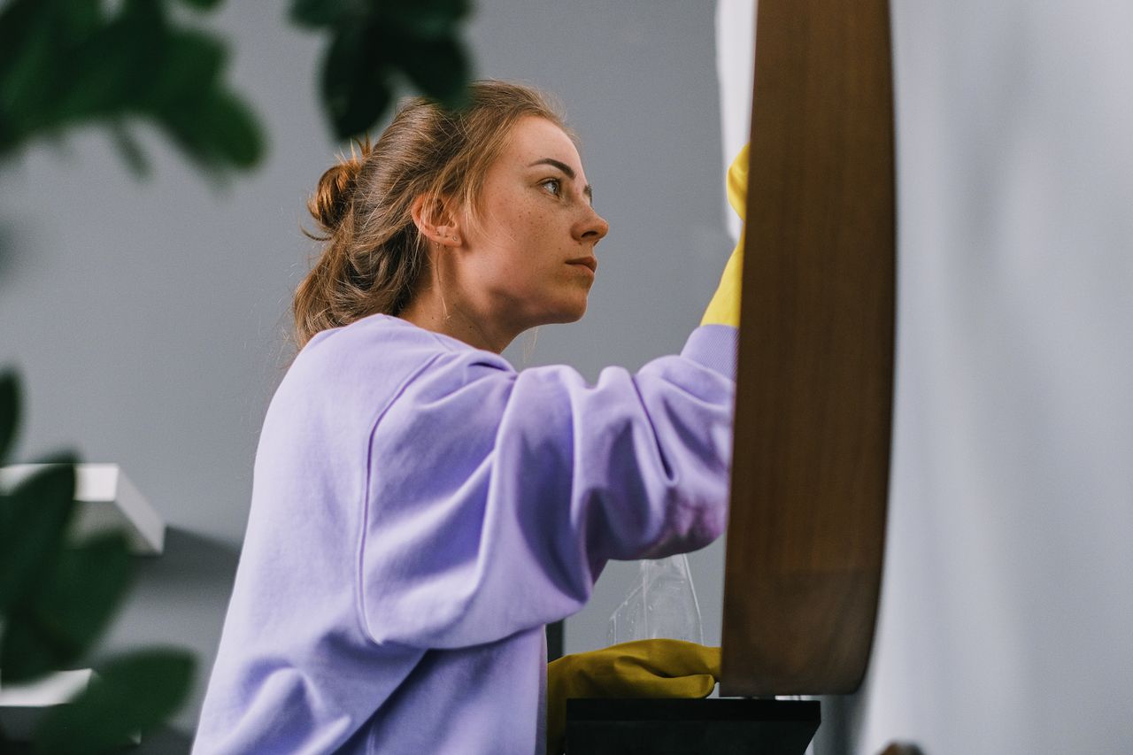 Profil latéral d'une jeune femme concentrée portant des gants en latex, debout en train de nettoyer un miroir dans une pièce lumineuse durant la journée